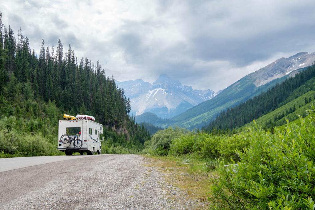 White RV parked on a mountain road, surrounded by lush green forests and distant snow-capped peaks.