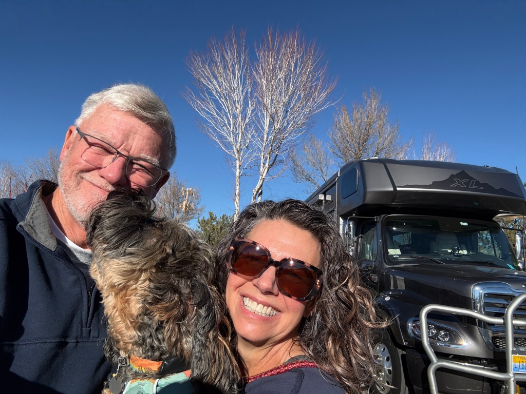 A smiling couple poses for a selfie with their small dog in front of a large RV under a clear blue sky.